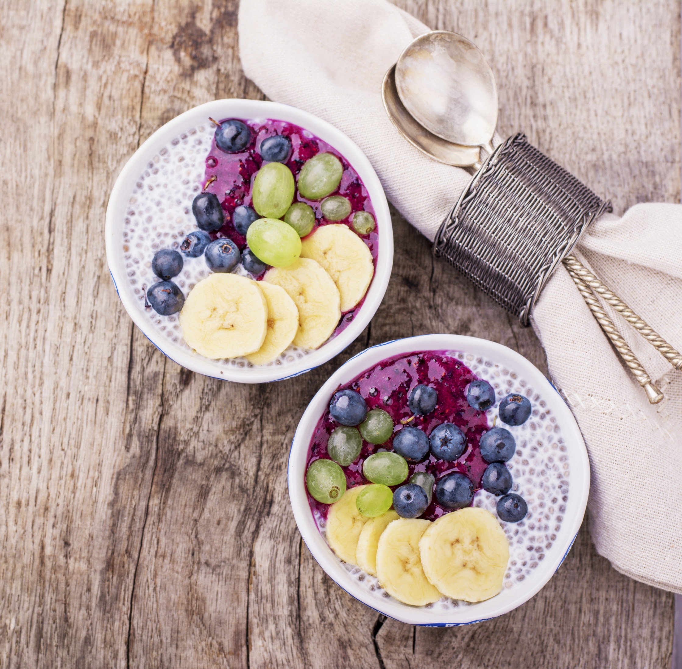 Chia Pudding for breakfast in bowls garnished with berry smoothies, grapes, petals of almond and blueberry in two portions on a wooden background with a napkin and spoon. Top view. selective Focus