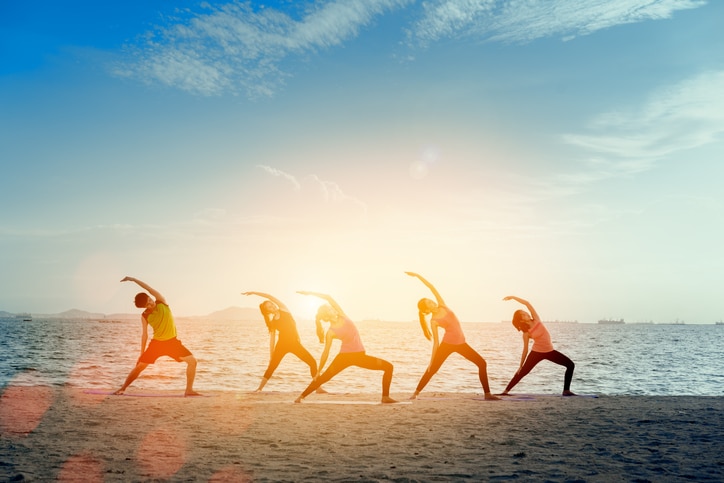 Young group man and woman yoga practice on mat relaxation in class exercise with pose fitness sport for healthy on the beach and seaside modern city at sunrise relax holiday vacation time in summer.