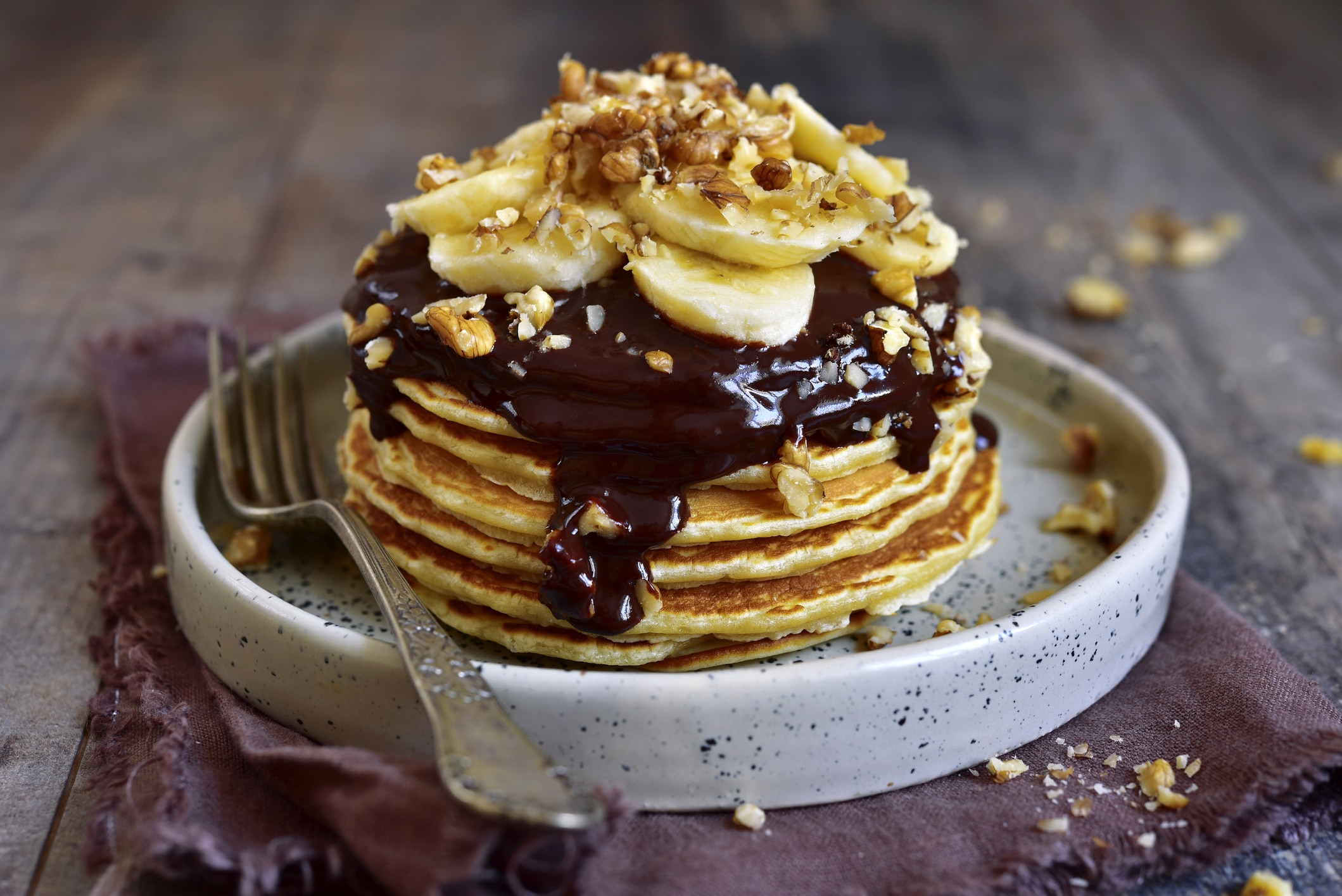 Stack of homemade delicious banana pancakes topped with chocolate sauce, banana slices and walnut on a plate on a dark wooden background.